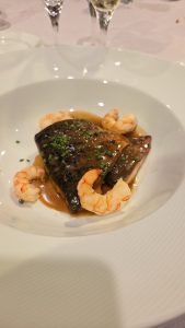 A seared fish fillet, likely sea bass or a similar white fish with a dark skin, garnished with fresh chives and served with peeled prawns in a savory reduction sauce.