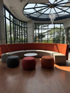 Interior shot of a bright modern lounge area with a curved sectional sofa featuring orange backrests and beige seating, paired with matching round ottomans, set against large grid windows and a dramatic circular glass skylight on the ceiling.
