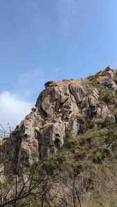 A rocky hillside with large boulders, dry grass, and sparse shrubs beneath a clear blue sky.
