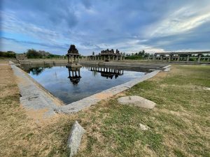 A wide view of a historic pond shows reflective water, granite steps, and rows of weathered pillars. The surrounding ruins and expansive cloudy sky create a peaceful scene in the ancient landscape. Location: Hampi, Karnataka