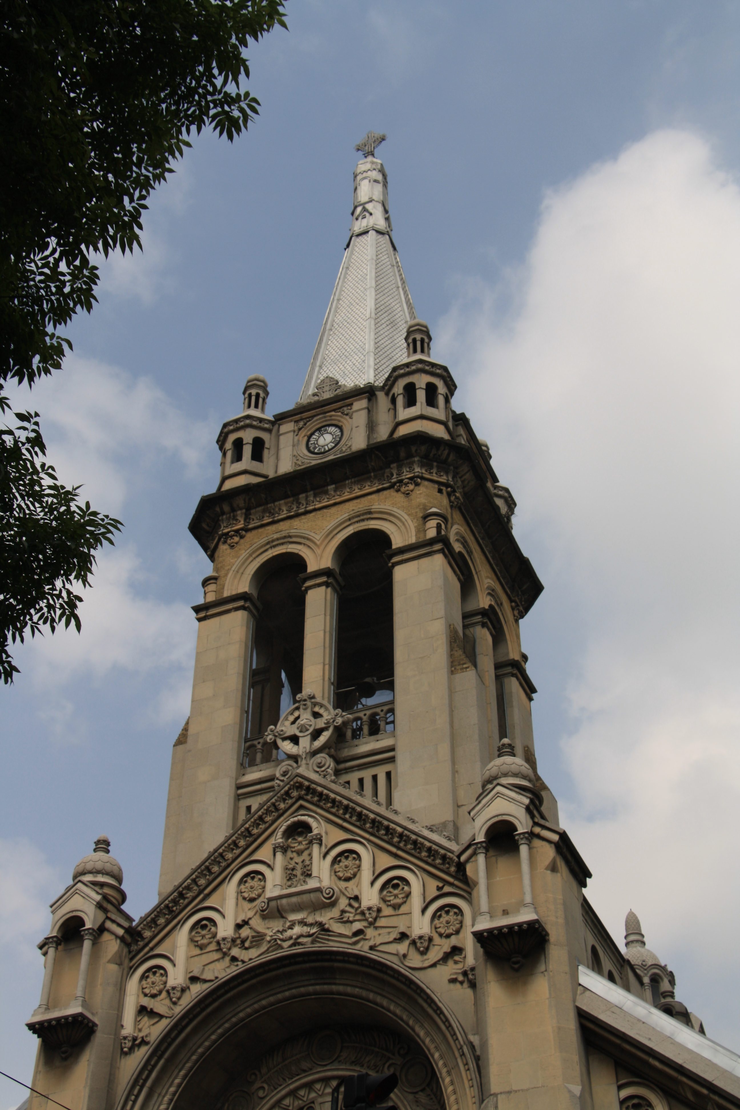 Low-angle shot of a tall Neo-Gothic church bell tower featuring intricate stone carvings, arches, a clock, and a pointed spire against a cloudy sky.