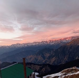 Pink and orange sunset clouds over snow-capped Himalayan mountain ridges, with a green-roofed building in the foreground. #wprajasthan
