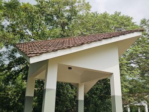 A small white pavilion with a traditional tiled roof stands among dense green trees. The simple architecture and natural surroundings create a peaceful scene. Located in Thonikadavu, Kozhikode, Kerala.