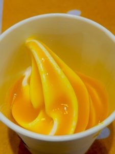 A close-up of bright yellow-orange soft-serve (mango) ice cream swirled into a white cup. Its glossy surface and vivid color stand out against the warm background. Taken at Palazhi, Kozhikode, Kerala.
