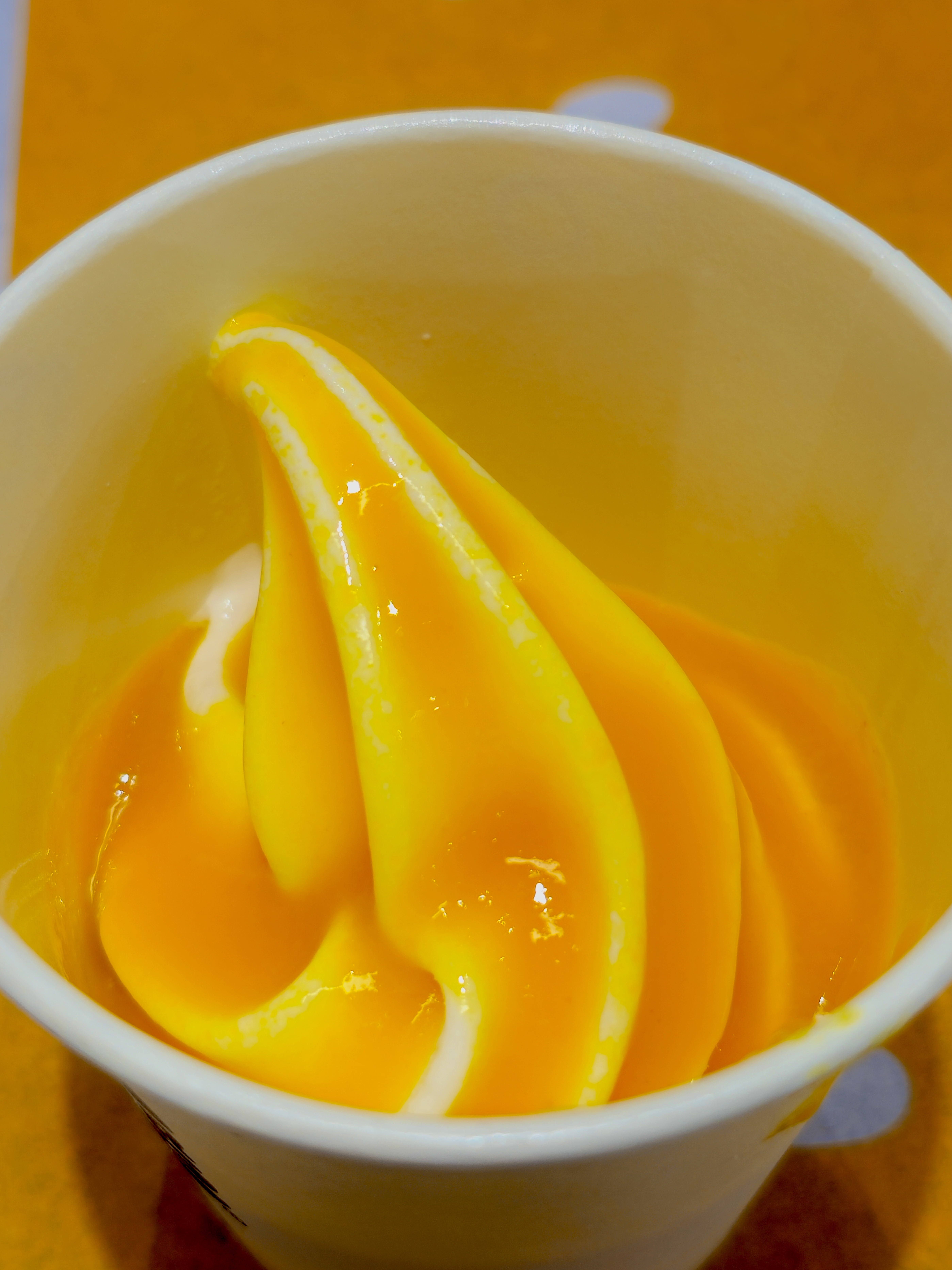 A close-up of bright yellow-orange soft-serve (mango) ice cream swirled into a white cup. Its glossy surface and vivid color stand out against the warm background. Taken at Palazhi, Kozhikode, Kerala.