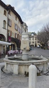 A stone fountain stands in a quaint square, featuring a tall, ornate column at its center topped by a majestic lion sculpture. Water flows from a lion's head design on the front of the pedestal, trickling down into the circular basin below. Surrounding the fountain, cobblestone paths lead to charming buildings with pastel-colored facades and shuttered windows. Some of the buildings have outdoor seating with striped tablecloths in front, giving the area a cozy, inviting atmosphere. Leafless trees and potted plants add to the scenery, while a partly cloudy sky looms above.