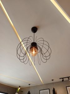 A modern pendant lamp featuring an open wireframe design of looping black metal petal wire shapes encasing a warm glowing filament bulb against a paneled ceiling.