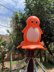 Red penguin-shaped playground ride in a garden with trees in the background

