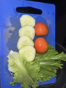 A blue cutting board displays several fresh ingredients. At the top, there are three thick slices of cucumber, their light green flesh visible, revealing tiny seeds inside. Below them, two medium-sized red tomatoes sit next to each other, their shiny skin reflecting light.