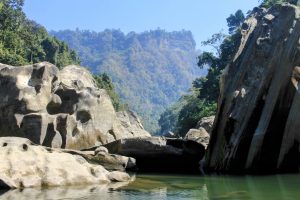Large pale grey boulders and a tall striped rock face beside calm green river water, with a forested hillside and blue sky in the background.
