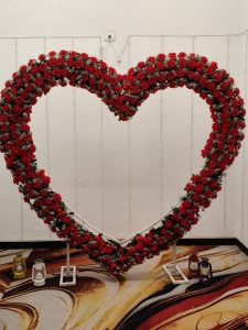 Large heart-shaped floral arch decorated with red roses, red flowers, and white accents, displayed indoors with lanterns at its base.