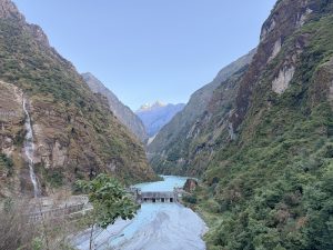 A hydroelectric dam structure set across a mountain river gorge, framed by steep, forest-covered cliffs and distant snow-capped peaks catching golden sunlight.