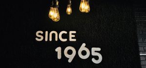 A black textured wall displays the words “Since 1965” beneath warm, glowing vintage light bulbs. The lighting creates a cozy, elegant atmosphere. Location: Ramanattukara, Kozhikode, Kerala.