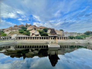An ancient pond with stone steps reflects a small pavilion, pillared structures, and rocky hills. The calm water mirrors the dramatic cloud-filled sky, creating a balanced composition of historic architecture and natural scenery. Location: Hampi, Karnataka.