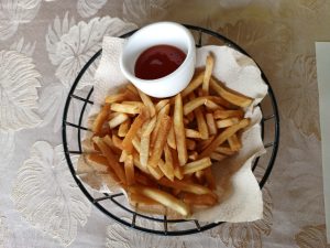 A wire basket lined with brown parchment paper holding a serving of golden French fries next to a small ceramic bowl of tomato ketchup.