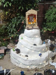 A festive white spiral mound decorated with glowing lights, a wooden temple with a colorful idol, marigold flowers, toy cars, and greenery.
