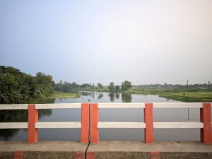 A red and white concrete bridge railing overlooking a calm rural river surrounded by green fields and trees beneath a pale cloudy sky.