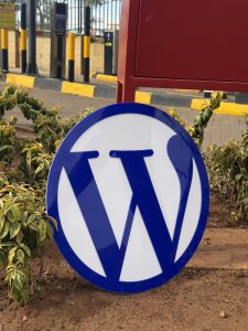 Blue circular WordPress logo with a white background placed on soil surrounded by small green bushes, with a paved area and yellow barriers in the background.
