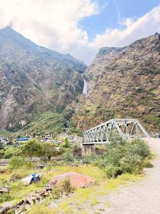 A grey steel truss bridge spanning across a stony riverbed, with lush green mountain slopes and a tall waterfall cascading in the background.