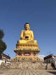 A large golden Buddha statue seated with a bowl in his lap, decorated robes, colorful prayer flags, and distant mountain peaks under a clear blue sky.