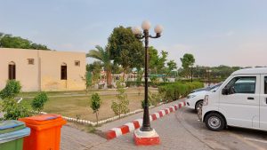 Parked white vans beside a landscaped garden with trees, hedges, streetlights, and colorful trash bins.