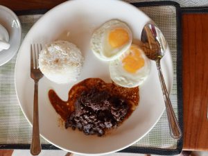 A plate featuring a deconstructed traditional Filipino cuisine breakfast made of tender beef tapa cooked in a dark savory glaze, accompanied by garlic rice and two sunny-side-up fried eggs.