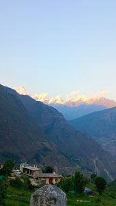 Snow-capped Himalayan mountains illuminated by golden sunlight above a green hillside village with houses and terraced slopes.