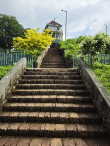 A stone staircase leads towards a round building, surrounded by lush greenery and bright yellow foliage under a partly cloudy sky.
