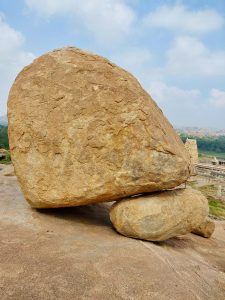 A massive, weathered boulder rests on smaller rocks near the historic temple complex of Hampi. The rocky foreground and distant ruins highlight the area's distinctive landscape. Location: Hampi, Karnataka