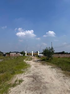 Daytime outdoor wide shot of a dirt track leading towards a white gated entrance with Bengali signage in a grassy field, under a bright blue sky with scattered white clouds.
