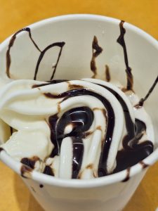 A close-up of vanilla soft-serve ice cream drizzled with chocolate sauce in a white cup. The swirls and contrasting colors create an appealing dessert photograph. Taken at Palazhi, Kozhikode, Kerala.
