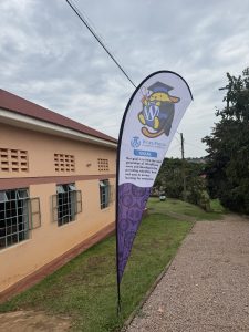 A WordPress Campus Connect feather banner stands on a gravel path beside a peach-coloured building with trees in the background.
