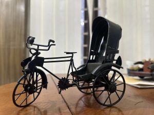 A small black metal model of a rickshaw sits on a wooden table, with a blurred indoor background featuring curtains and papers. 
