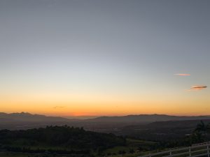 Sunset over mountains and trees with a white fence in the foreground.