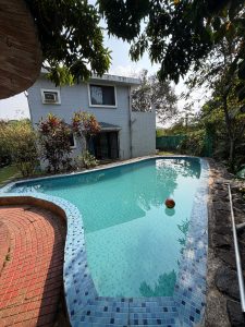 A curved turquoise swimming pool surrounded by tropical greenery, with a two-story villa in the background and tree branches overhead.
