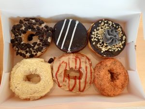 An open cardboard bakery box filled with six assorted gourmet donuts featuring varied toppings of crushed cookies, chocolate icing, chopped nuts, and flavored glazes.