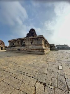 An ancient stone temple stands in a spacious courtyard paved with weathered granite slabs. Intricate architectural details and a partly ruined tower rise beneath a cloudy blue sky. Location: Hampi, Karnataka.
