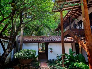 A white house with tiled roofs and white walls, the house has an opening in the middle that works as a door, stairs on the side, a tree with fruit, and green vegetation.