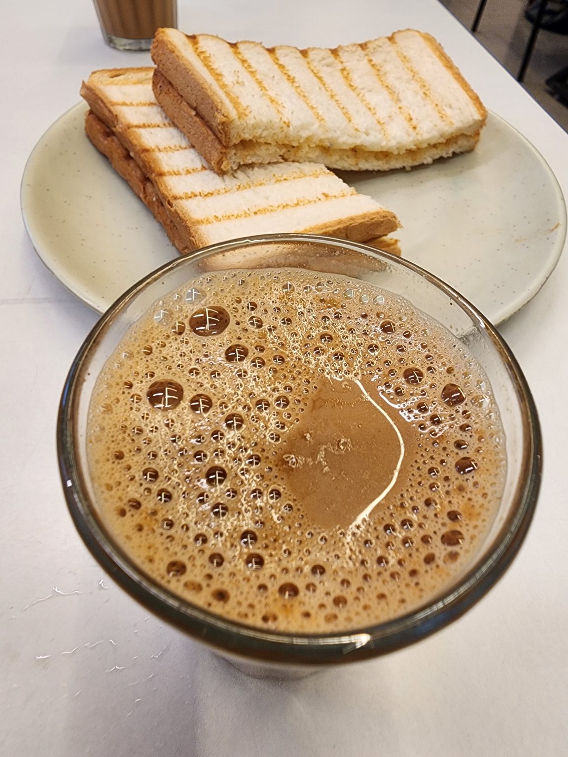 A glass filled with frothy, dark brown liquid, likely a coffee or chocolate drink, is placed prominently in the foreground. In the background, there’s a light-colored round plate holding several slices of grilled bread, which have a golden-brown crust and are arranged neatly. The slices appear to have been toasted with distinct grill marks, and some have been partially cut, revealing a soft, white interior. A blurred cup of similar drink is visible behind the plate, adding to the café setting. The surface beneath the items is white, creating a clean, simple backdrop.