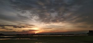 A peaceful sunset over a calm riverside landscape beneath dramatic clouds.