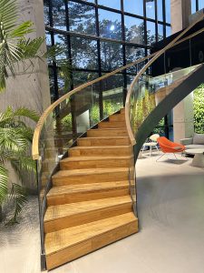 A modern interior space features a gracefully curved wooden staircase with a sleek glass railing, leading upward.