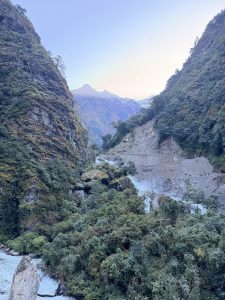 A deep mountain valley framed by steep, forest-covered cliffs and a gravelly landslide area, with a rushing glacier river at the canyon floor leading toward snow-dusted distant peaks