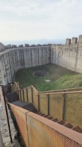 The image shows a large, circular stone structure resembling the interior of an ancient fortress or castle. Surrounding the circular area are high stone walls with a rough texture, made of gray stones. There are remnants of circular structures and metal poles set in the ground, suggesting a previously used space. A set of metal steps, framed with rust-colored railings, descends into the center of this structure. The area is mostly green, with patches of grass visible. The sky above is light blue with scattered clouds, and distant mountains can be seen on the horizon. The overall atmosphere gives a sense of history and age, with an open, airy feel created by the high walls and expansive exterior view.
