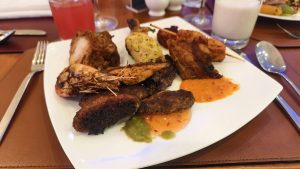 A white plate filled with mixed grilled items like chicken kebab, fried fish, and prawns served with spicy sauces on a dining table.