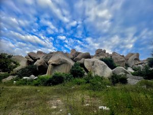 Massive granite boulders form a rugged, rocky landscape dotted with green shrubs and grass. Dramatic clouds spread across the vivid blue sky, highlighting this historic region's distinctive geology in the evening. Location: Hampi, Karnataka.