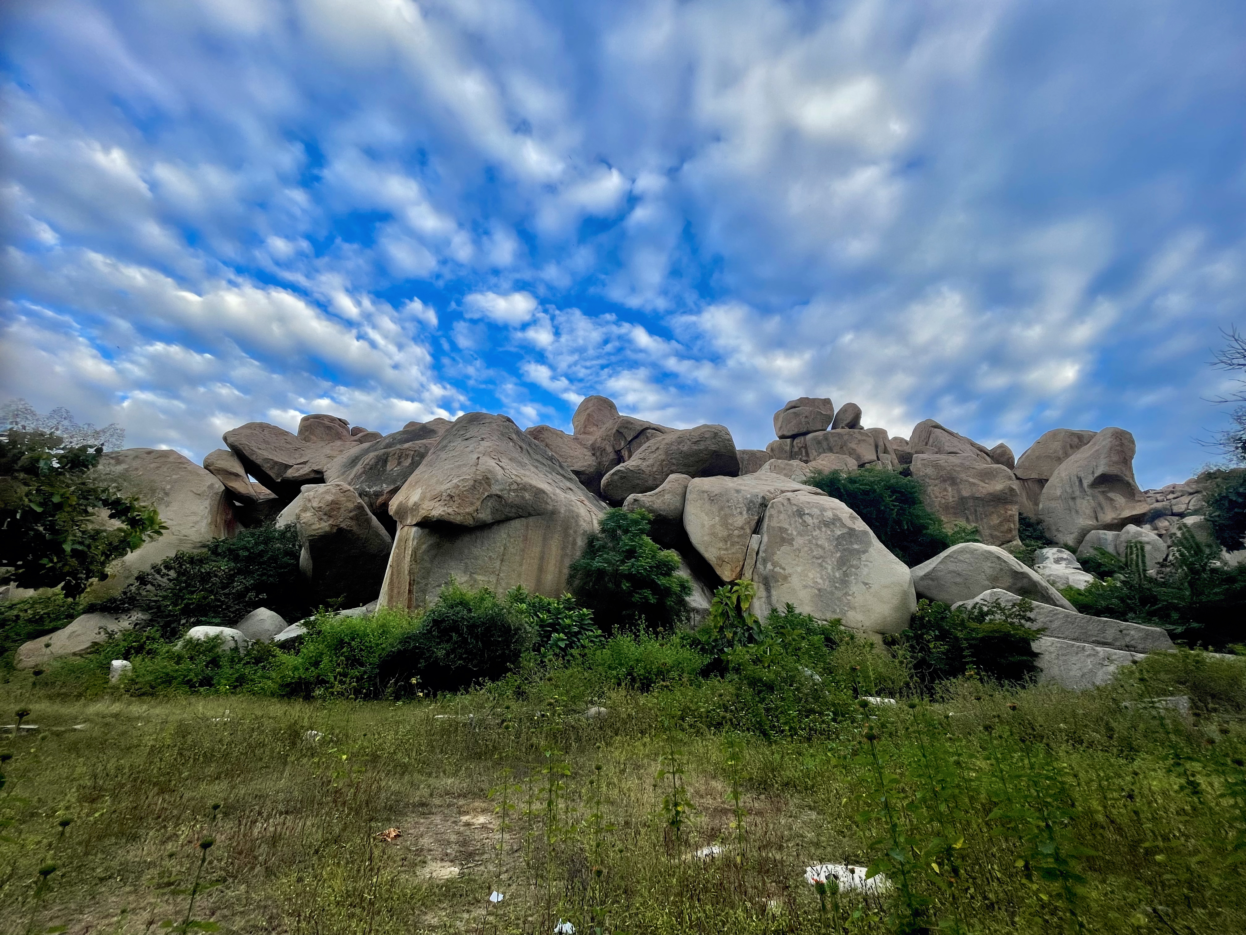 Massive granite boulders form a rugged, rocky landscape dotted with green shrubs and grass. Dramatic clouds spread across the vivid blue sky, highlighting this historic region's distinctive geology in the evening. Location: Hampi, Karnataka.