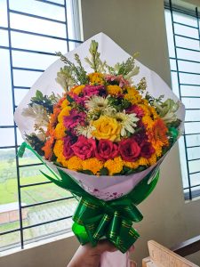 A vibrant flower bouquet featuring red and yellow roses, gerbera daisies, and marigolds wrapped in white paper with a green ribbon.
