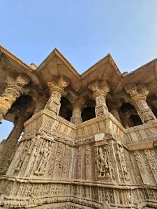 A dramatic low-angle photograph captures the richly decorated pillars, sculpted figures, and geometric stonework of the Sun Temple. Warm sunlight enhances the intricate craftsmanship. Location: Sun Temple, Modhera, Mehsana district, Gujarat.
