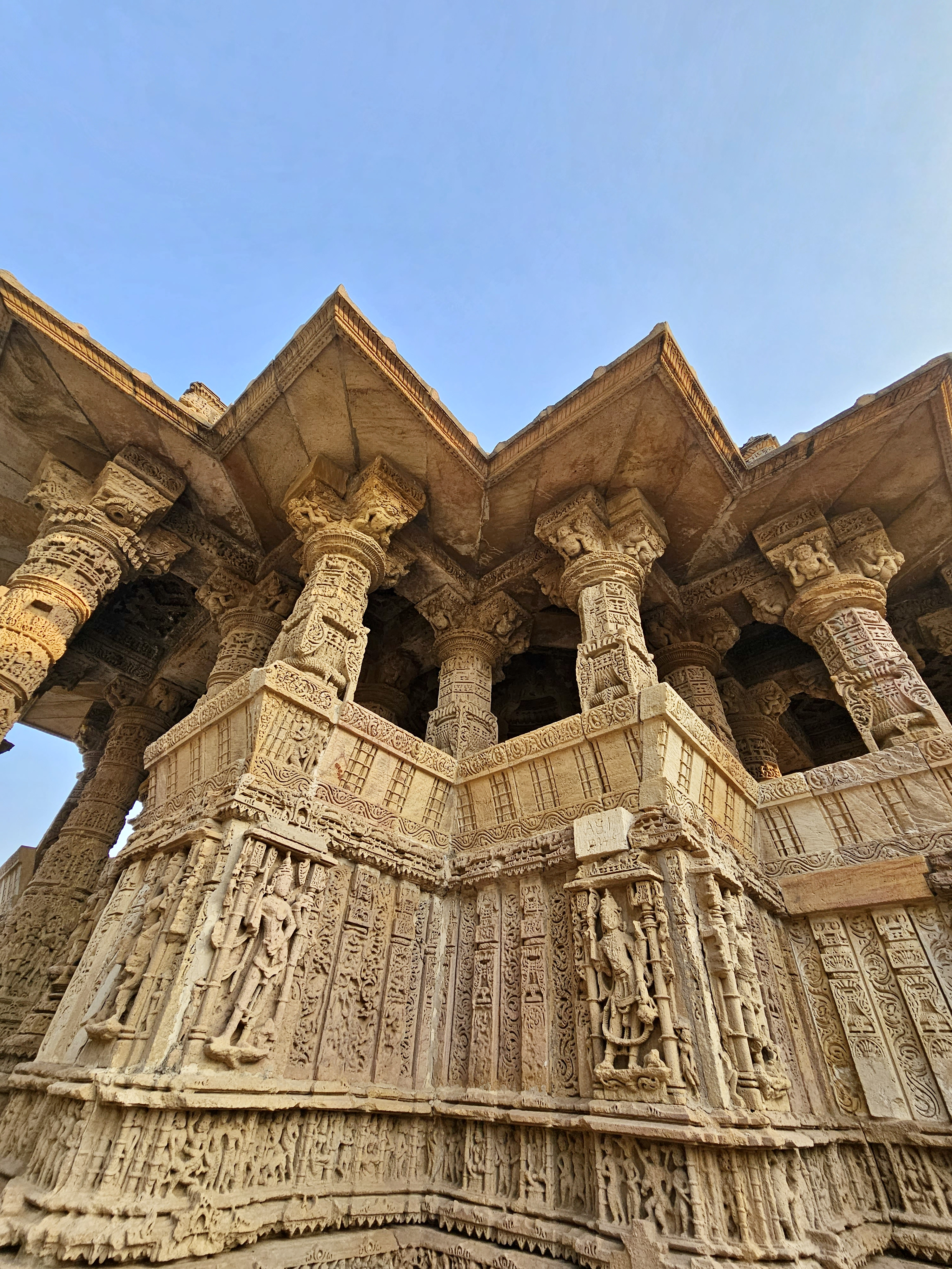 A dramatic low-angle photograph captures the richly decorated pillars, sculpted figures, and geometric stonework of the Sun Temple. Warm sunlight enhances the intricate craftsmanship. Location: Sun Temple, Modhera, Mehsana district, Gujarat.