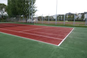 The image shows a tennis court with a red playing surface, outlined by white lines that define the court’s boundaries. There is a black net in the center, stretching across the court. On one side, the surface transitions to a green area.

